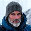 강인한 표정의 베테랑 등반가가 얼음 절벽 앞에서 카메라를 응시하는 모습.